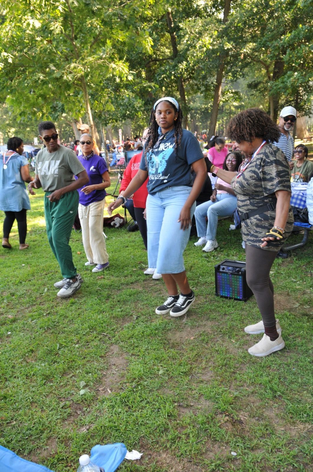People dancing outdoors on a grassy field at a park, with trees and others watching nearby.