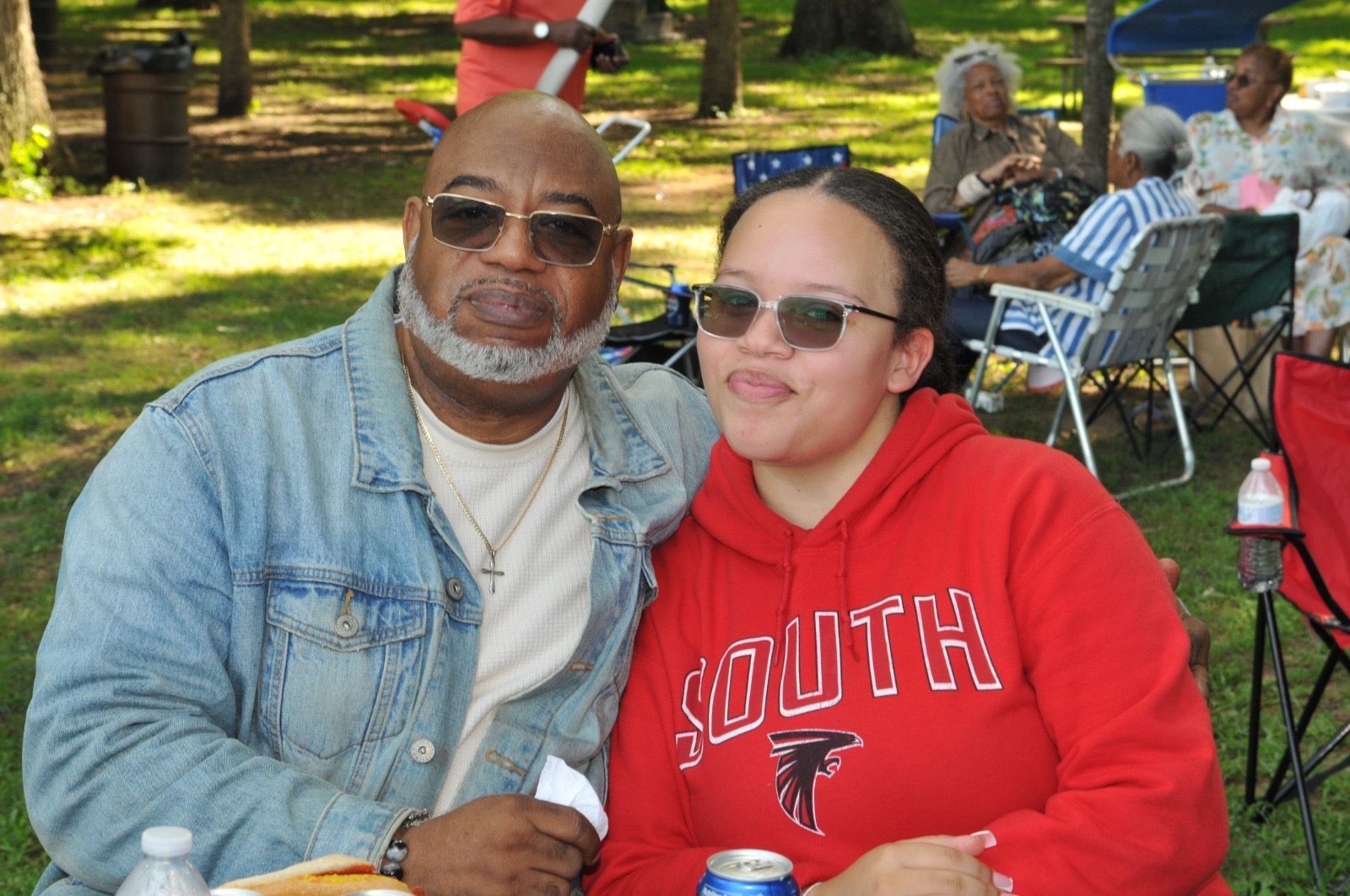 Two people seated outdoors on grass, smiling in denim jacket and red hoodie at a park picnic.