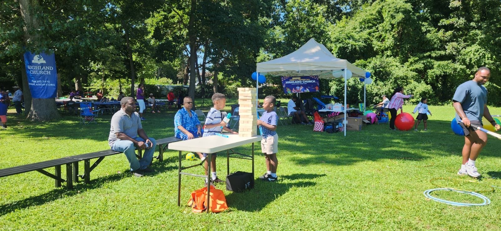 People sitting at picnic tables in a sunny park with trees and a canopy tent.