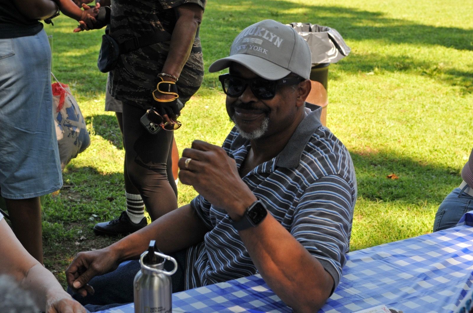Man seated at a picnic table outdoors, smiling and holding a drink, with a blue checkered tablecloth nearby.