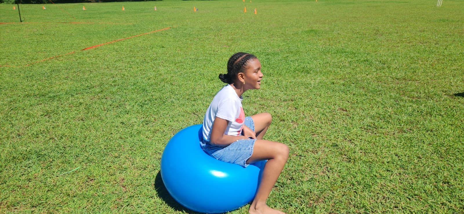 Person sitting on a blue inflatable ball in a grassy field, facing right and smiling.