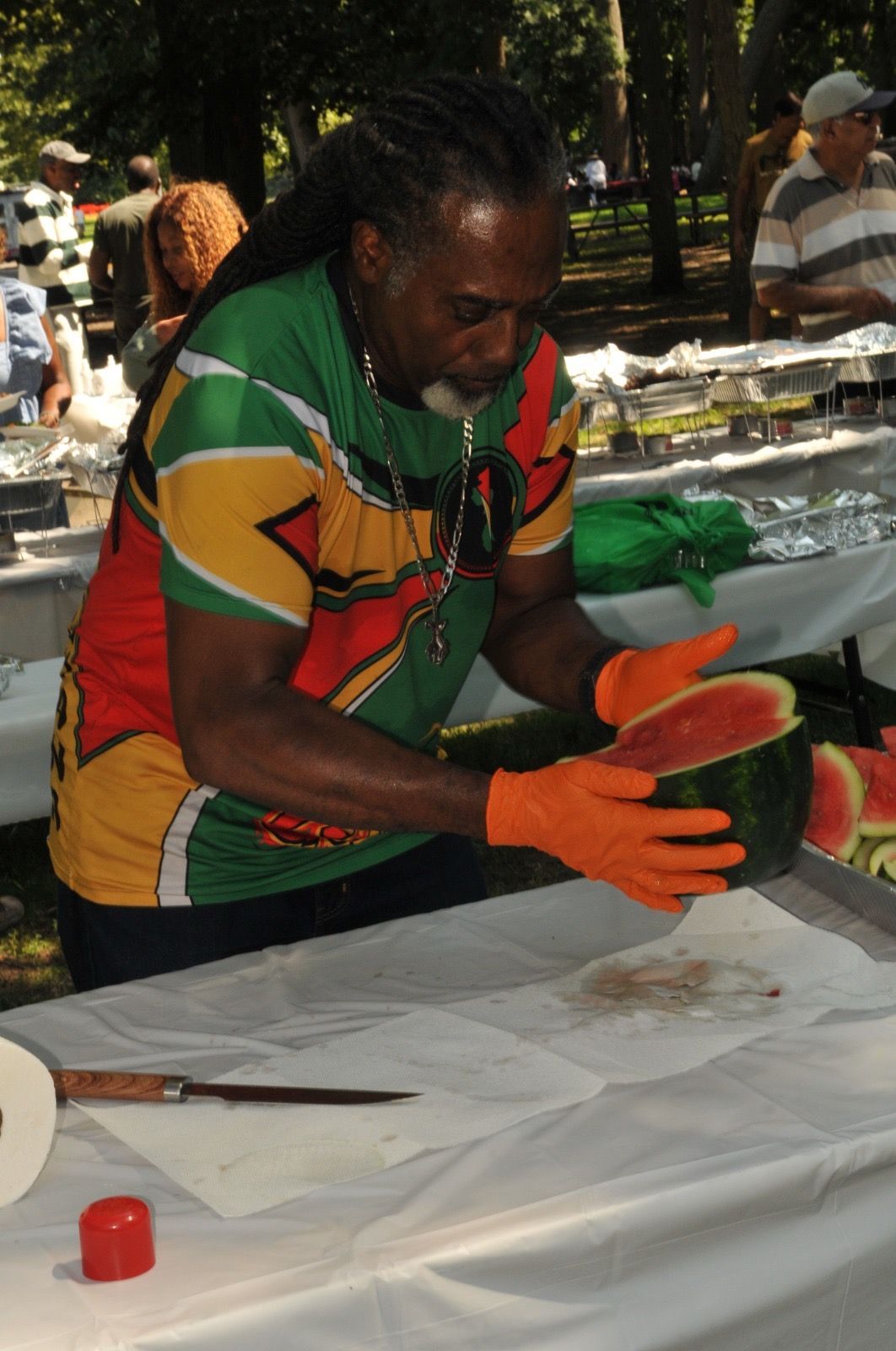 Person in a colorful shirt and orange gloves slicing watermelon at an outdoor table.