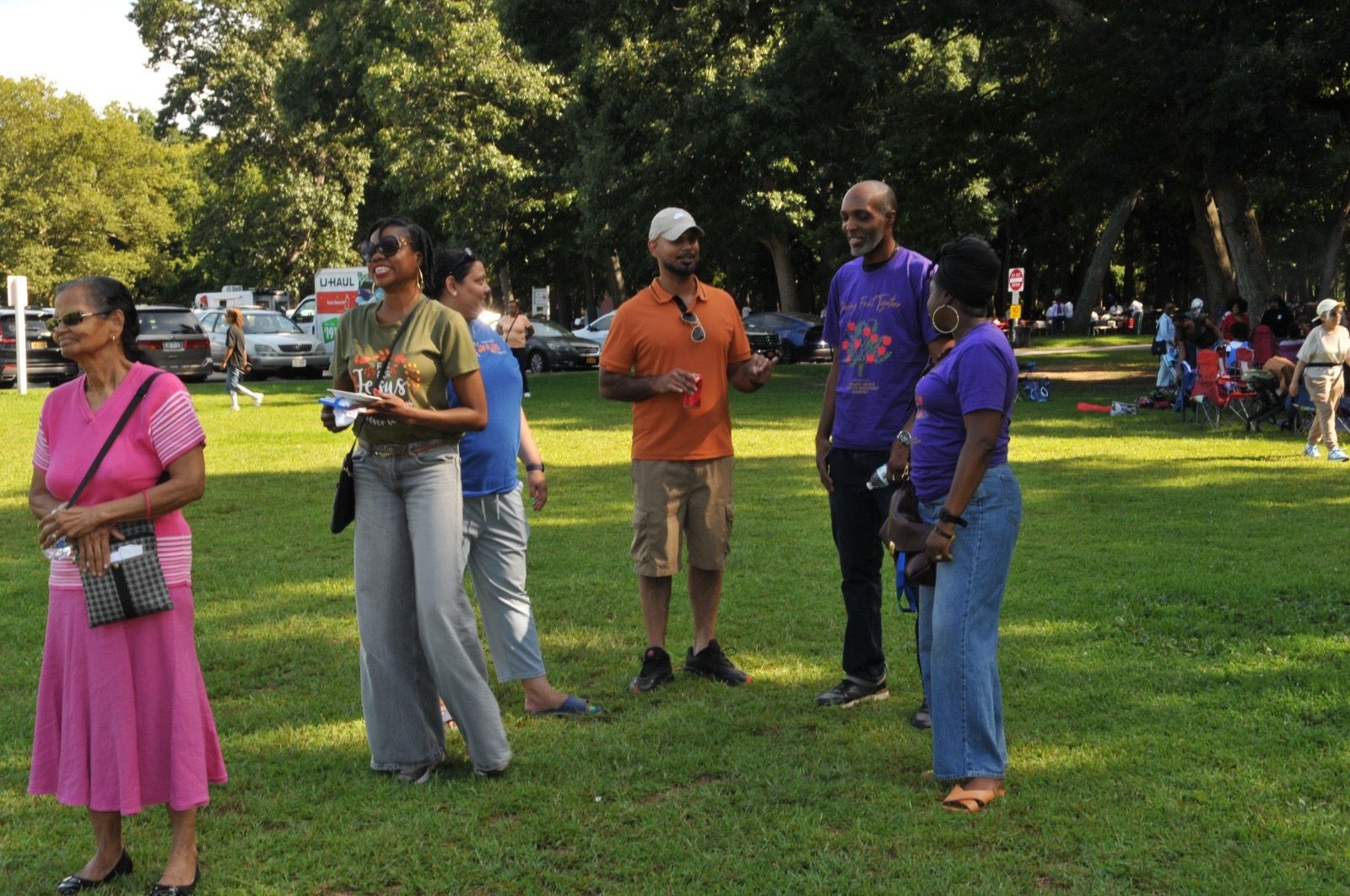 People standing and chatting in a sunny park, with trees and more people in the background.