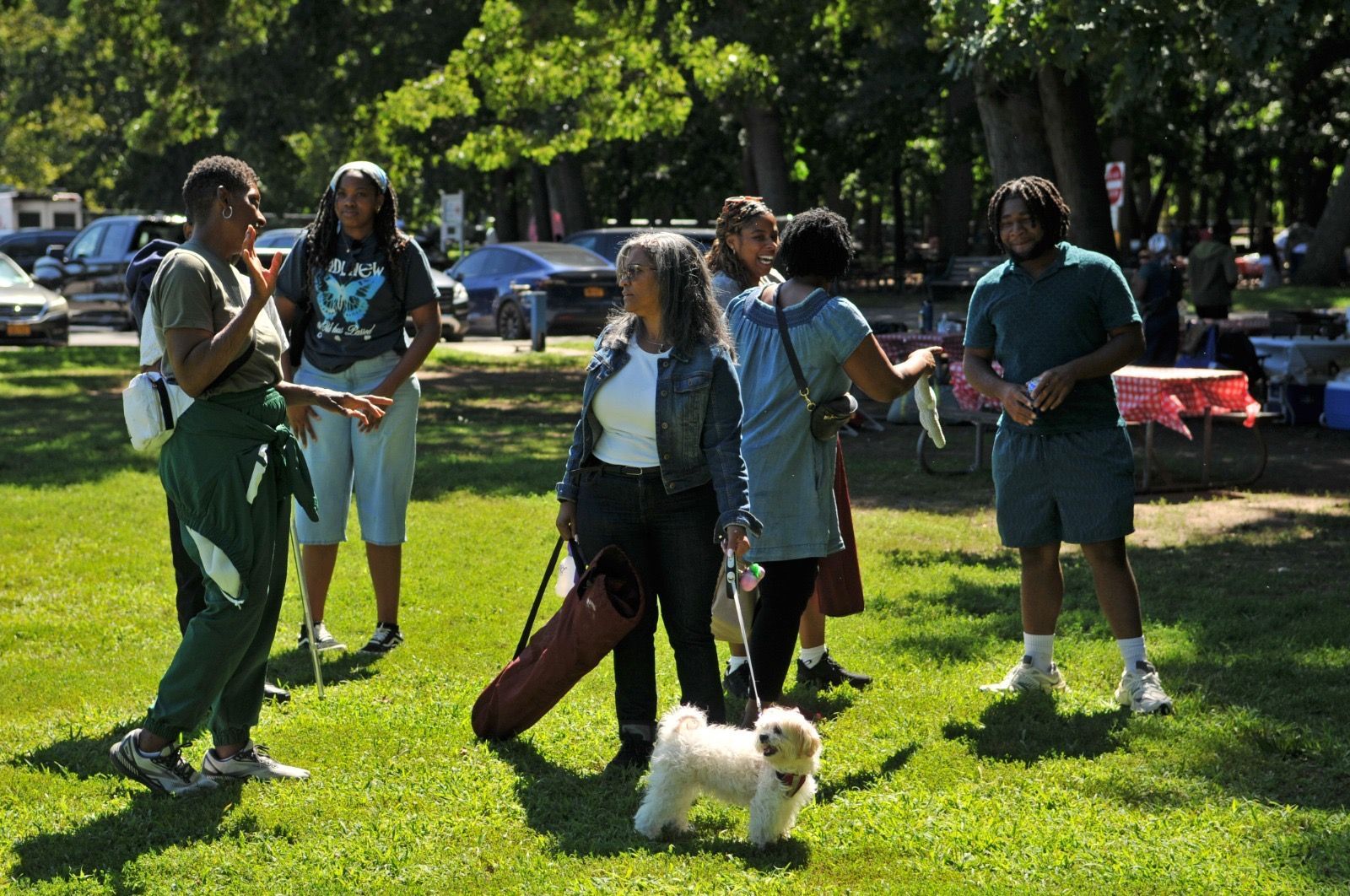 People standing on a sunny lawn, one holding a guitar case, with a small white dog in front