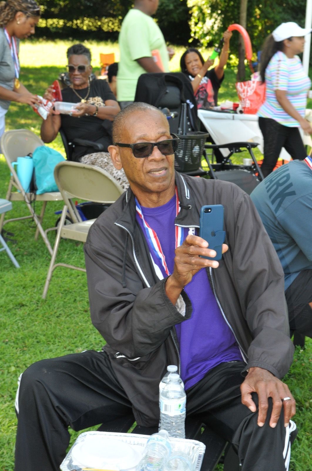 Man seated outdoors on folding chair, holding a phone, with water bottle in front and people in the background