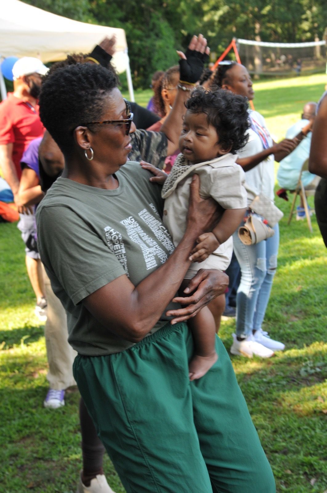 Woman holding a child at an outdoor gathering in a park, with people and tents in the background