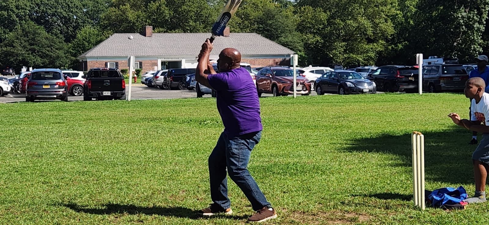 Cricket player in purple swings bat on a grassy field while a wicketkeeper crouches nearby.