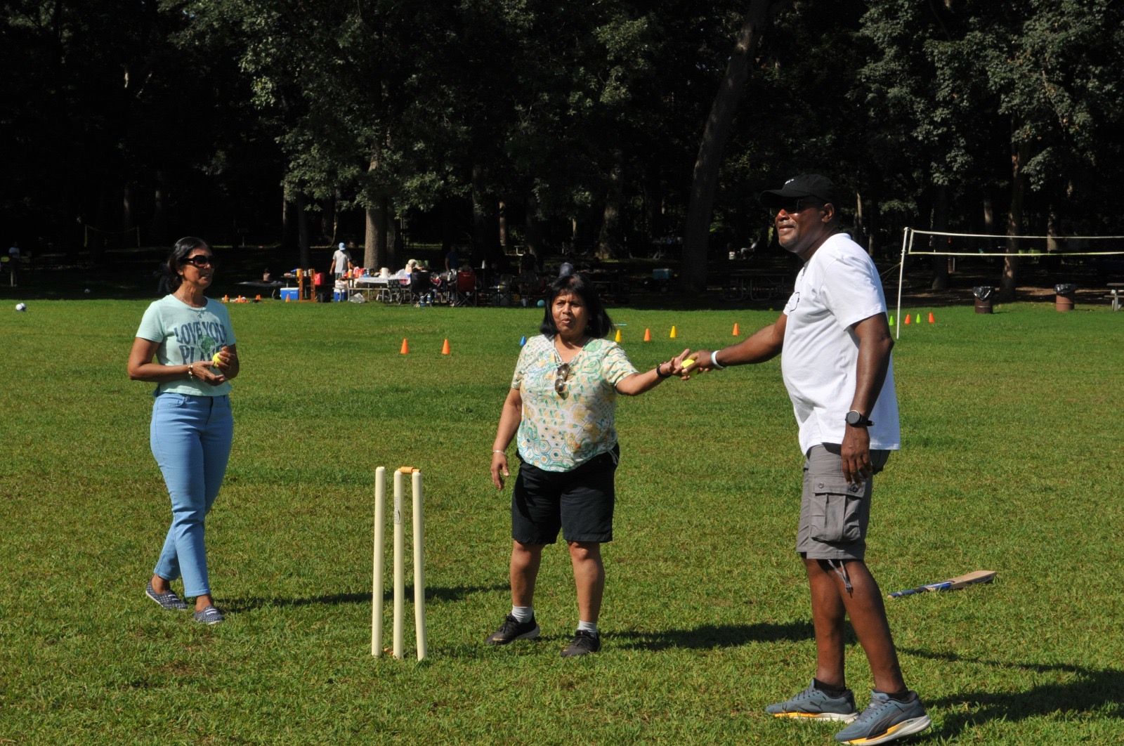 Three people on a grassy field playing cricket; one person holds the ball near the stumps.