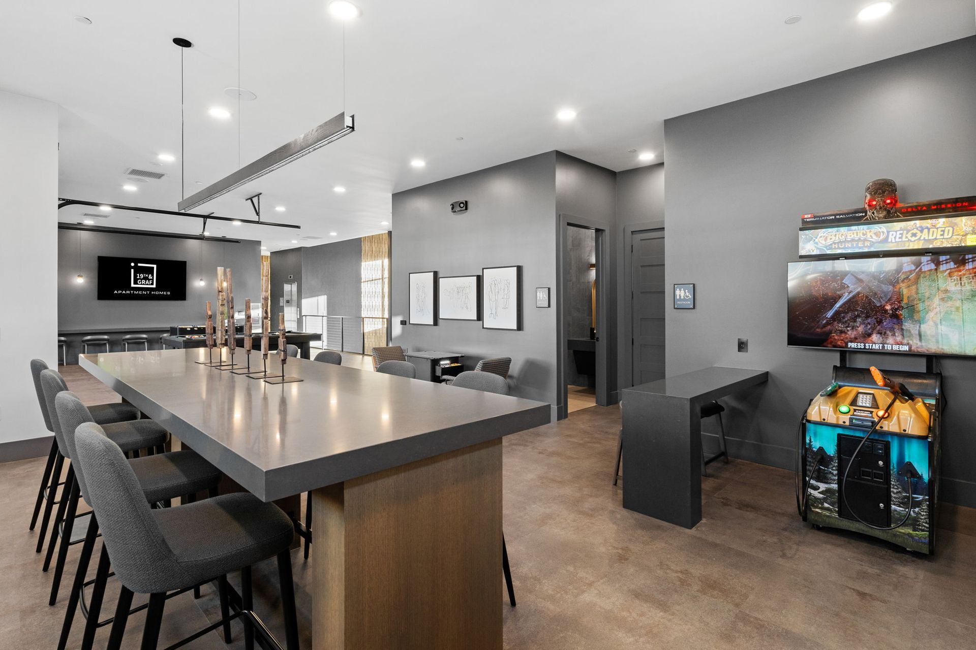 Modern lounge with long bar, stools, arcade game, gray walls, and recessed lighting.