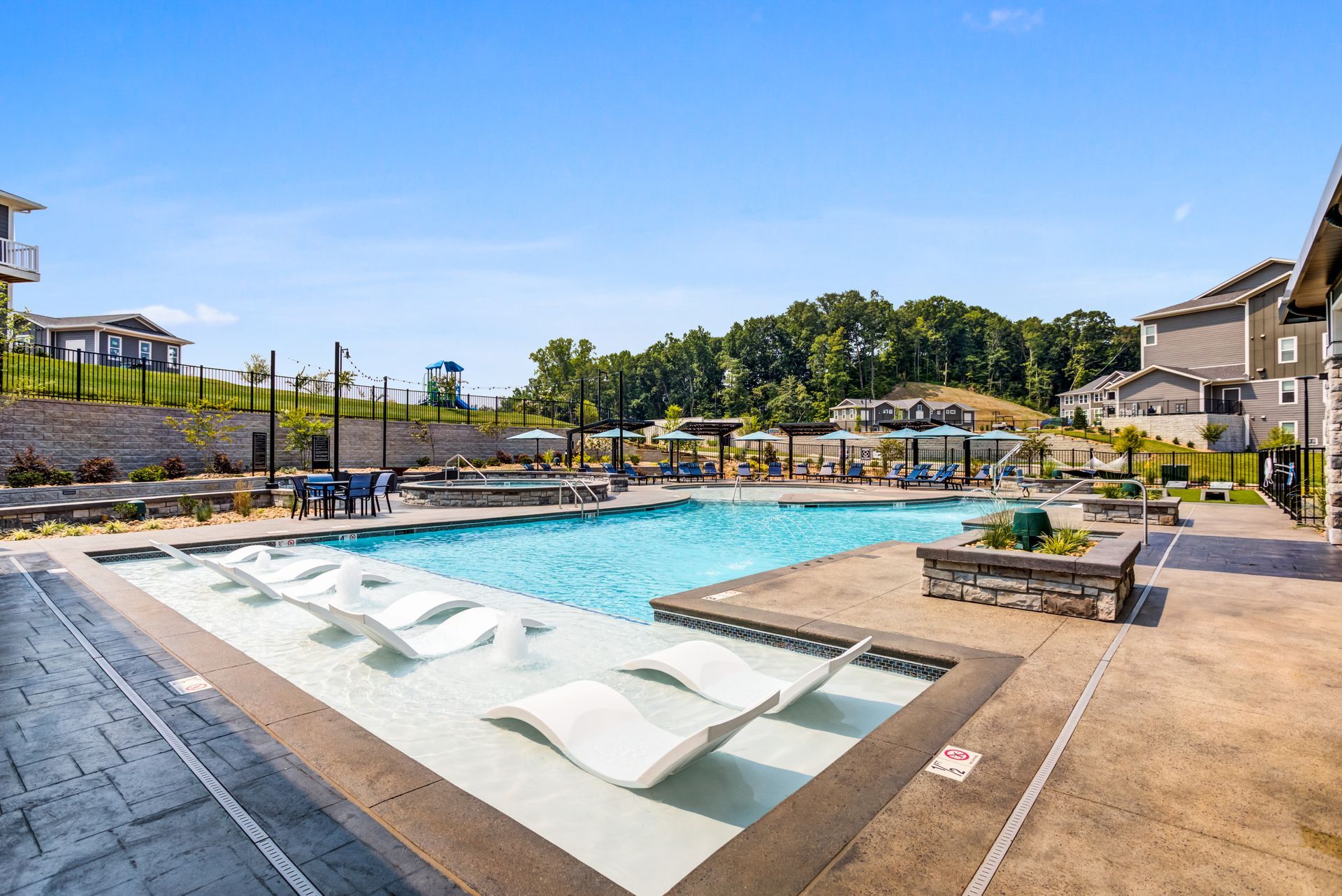 A community swimming pool with lounge chairs, blue water, and surrounding buildings under a sunny sky.
