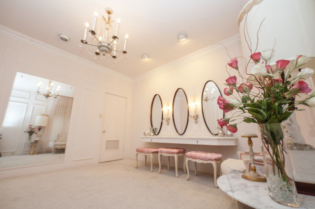 A bright, elegant dressing room with three oval vanity mirrors, light pink stools, a crystal chandelier, and floral decor.
