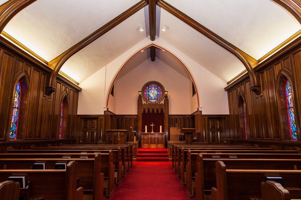 The interior of a church with wooden pews, a red carpeted aisle, paneled walls, and stained glass windows.