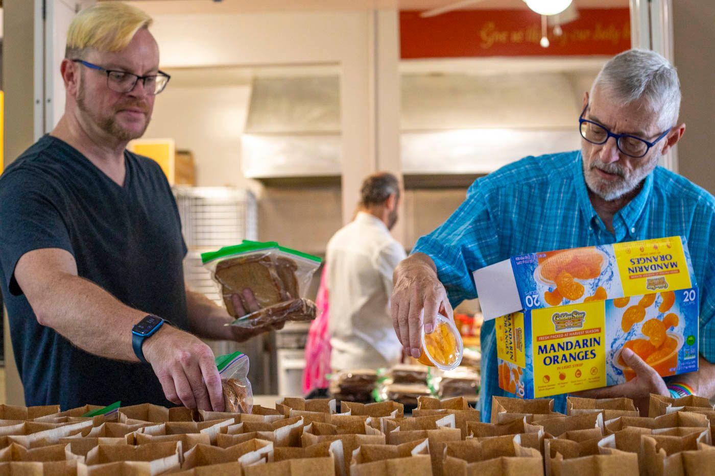 Two men pack brown paper bags with food in a kitchen; other people are present in the background.