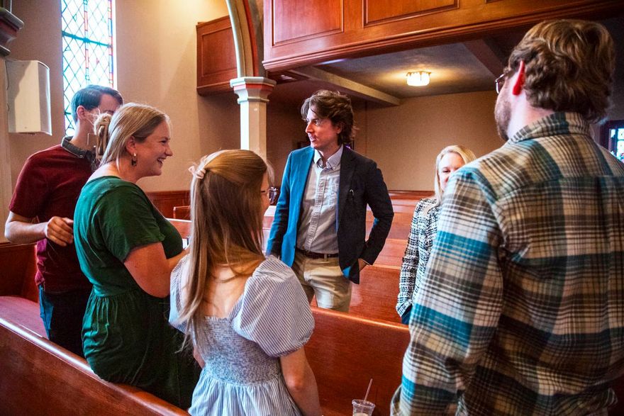 Group of people in a church setting, talking. Interior shot with pews and stained glass.