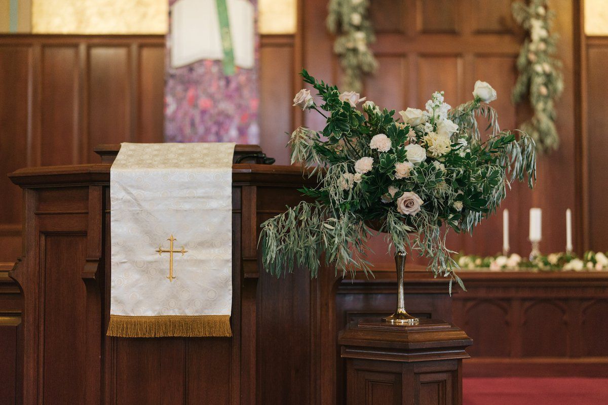 A wooden church pulpit draped with a white paraments cloth, featuring a golden cross, next to a large floral arrangement.