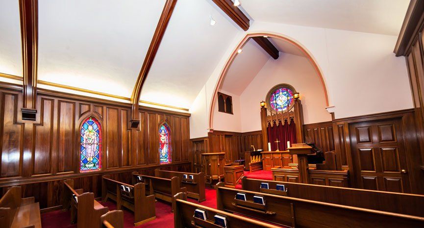 A sanctuary with wood-paneled walls, pews, stained glass windows, and a red carpet leading toward a central altar area.
