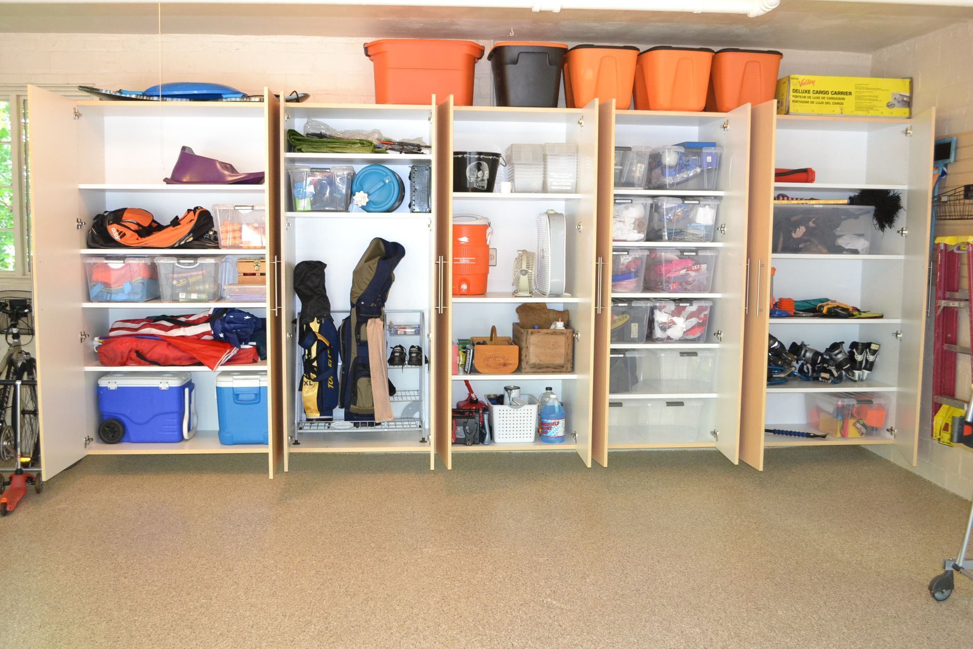 Open wall-mounted garage cabinets with organized bins, coolers, and sports gear over a tan flake flo