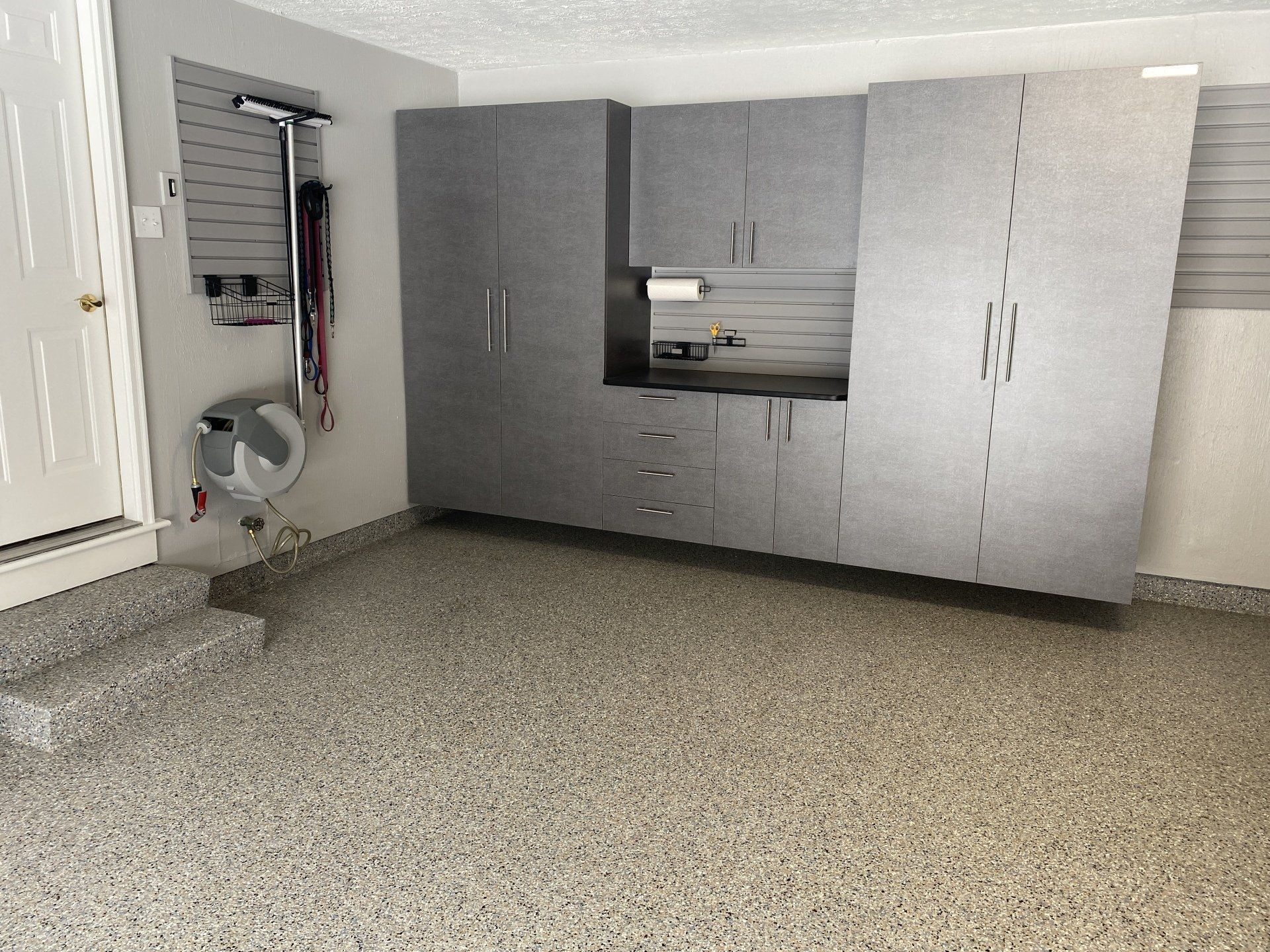 Gray wall-mounted garage cabinets with slatwall, hose reel, and tan flake polyaspartic floor in a Cl