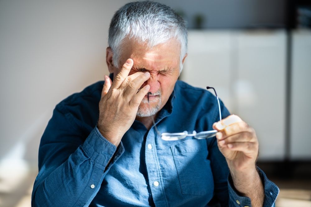 An older man is rubbing his eyes while holding a pair of glasses.