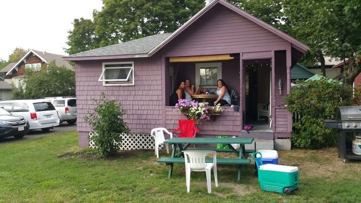 Purple cottage with people on a porch, green picnic table, and cars in a grassy yard.