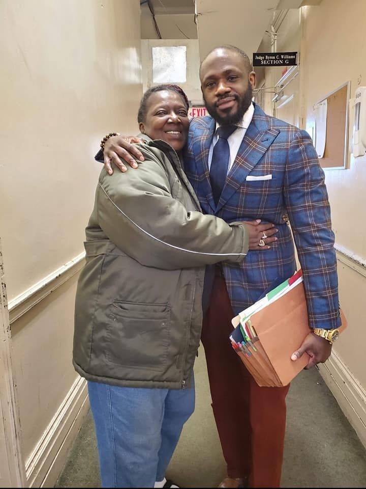 Two people smiling and hugging in an office hallway; one person wears a blue plaid blazer and holds a document folder.