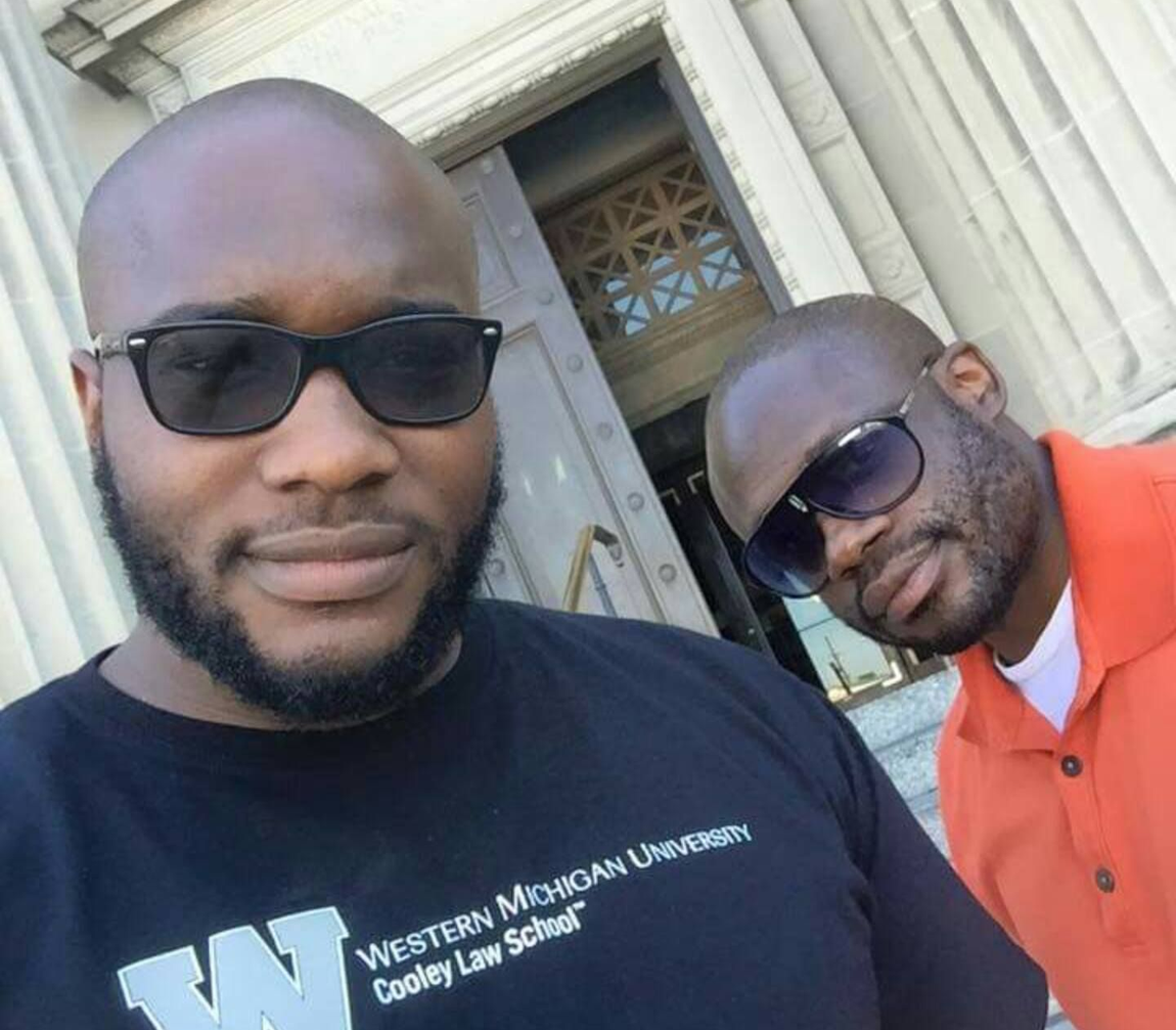 Two men wearing sunglasses stand in front of a building entrance; one wears a black university shirt, the other an orange polo.