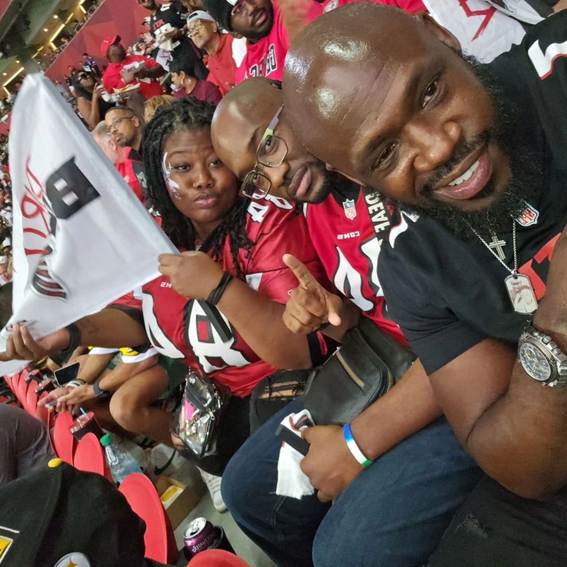 A group of people wearing football jerseys and smiling while seated in a stadium, one holding a team pennant.