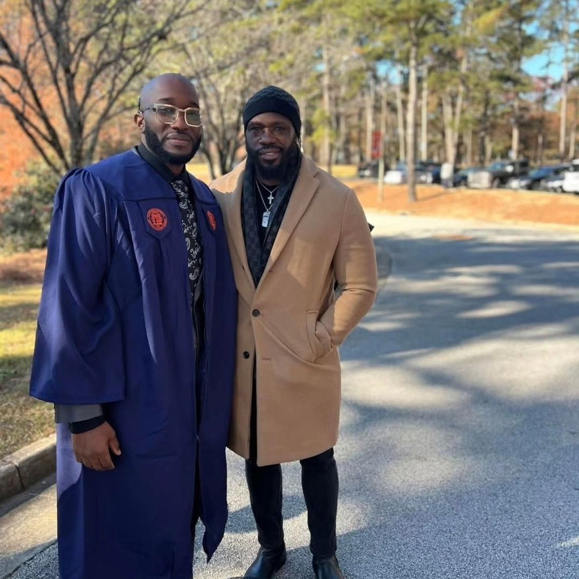 Two people standing side-by-side outdoors; one wears a graduation gown, the other a camel coat, standing on a paved lot.
