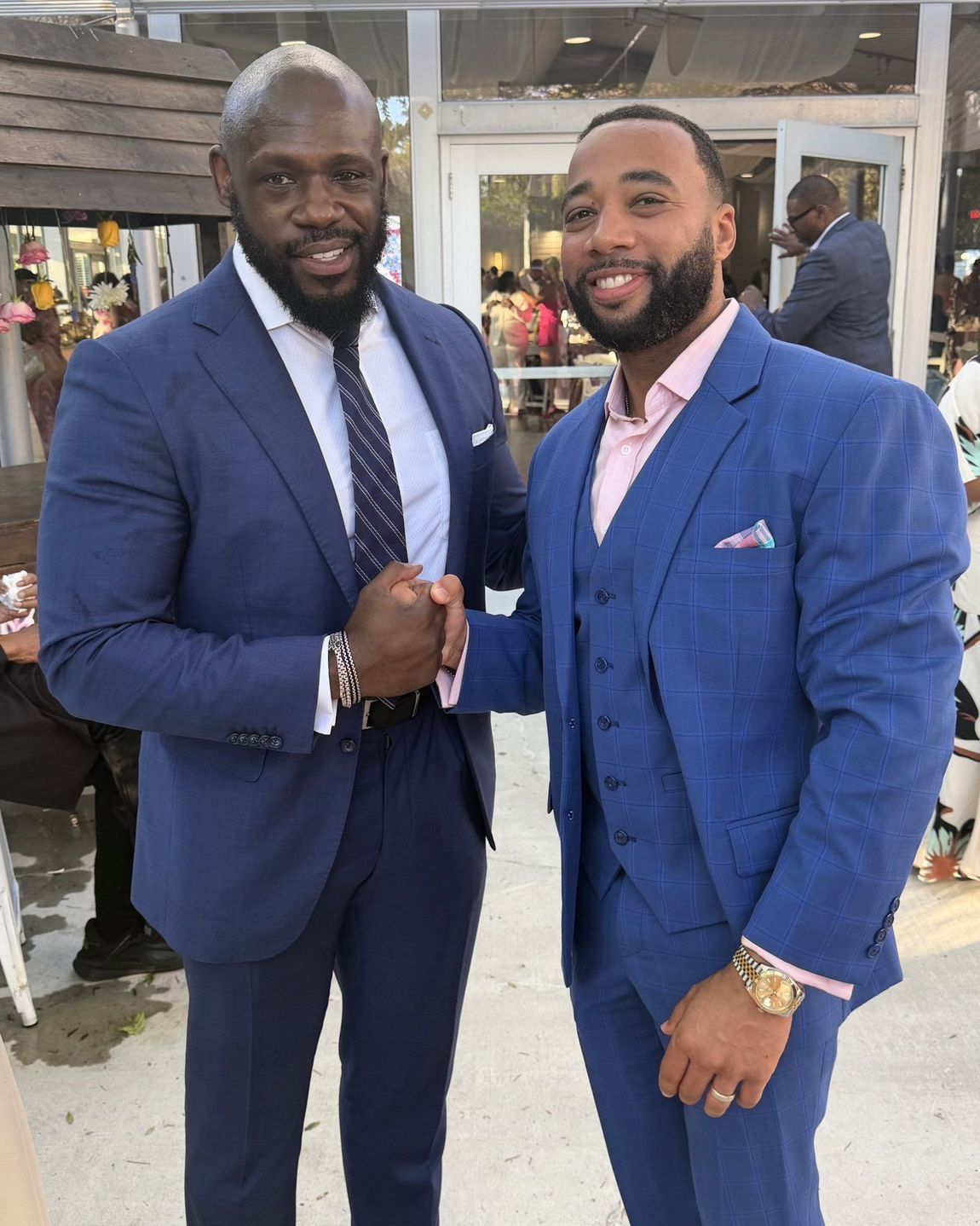 Two men in blue suits shaking hands while smiling in an outdoor setting with blurred figures in the background.