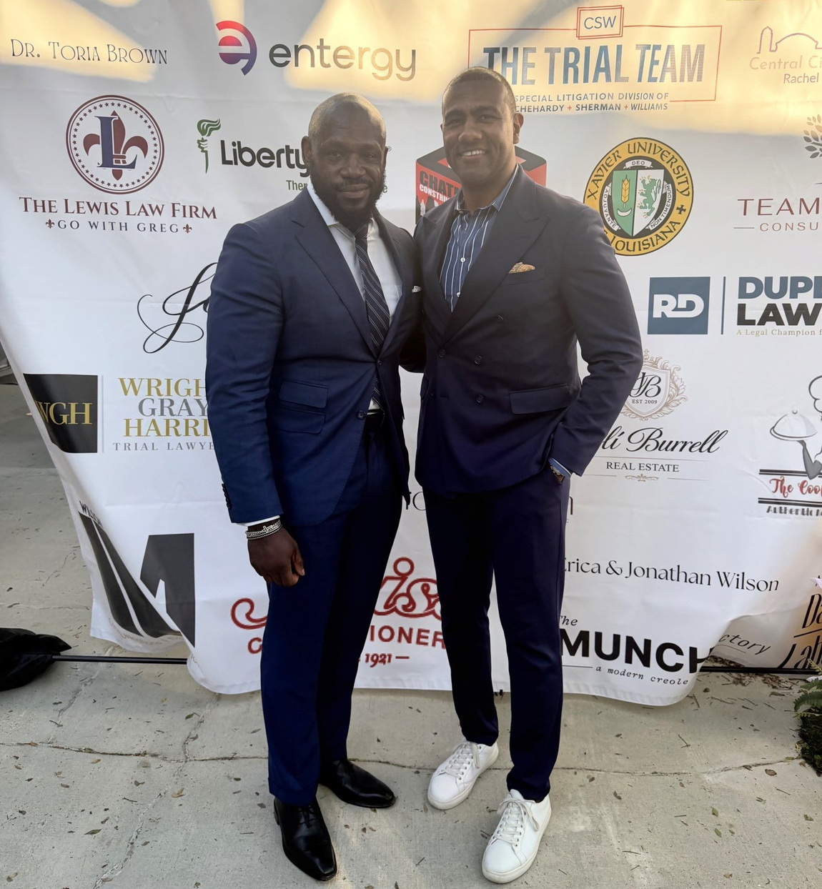 Two men in dark blue suits pose for a photo in front of a branded event backdrop featuring logos of various law firms.