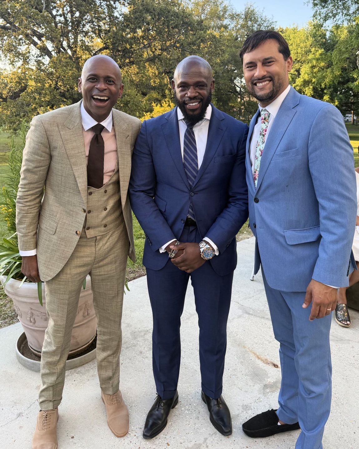 Three men in suits—tan, blue, and light blue—standing together outdoors smiling in front of a tree-lined backdrop.
