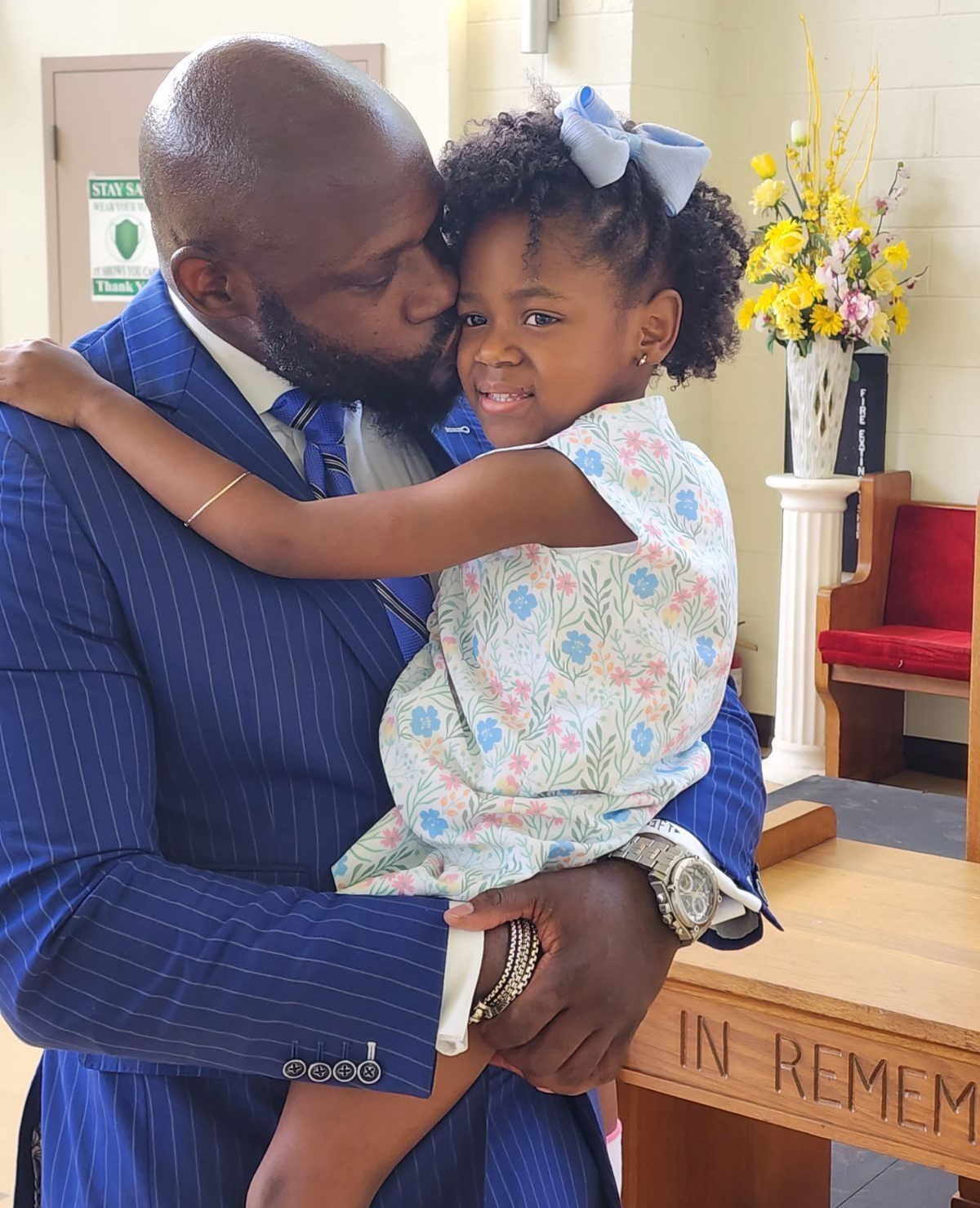 A person in a blue pinstripe suit kisses a child wearing a floral dress and blue hair bow in a church.
