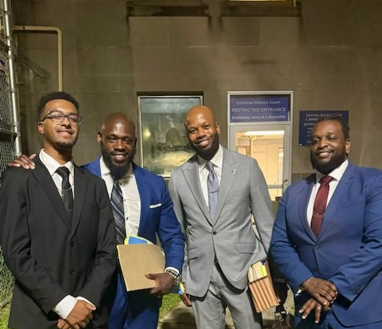 Four people in business suits stand together smiling outside a building entrance at night.