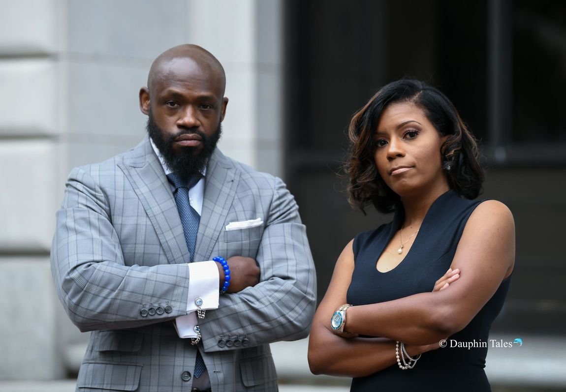 A man in a grey suit and a woman in a black top standing side-by-side with arms crossed, looking at the camera.