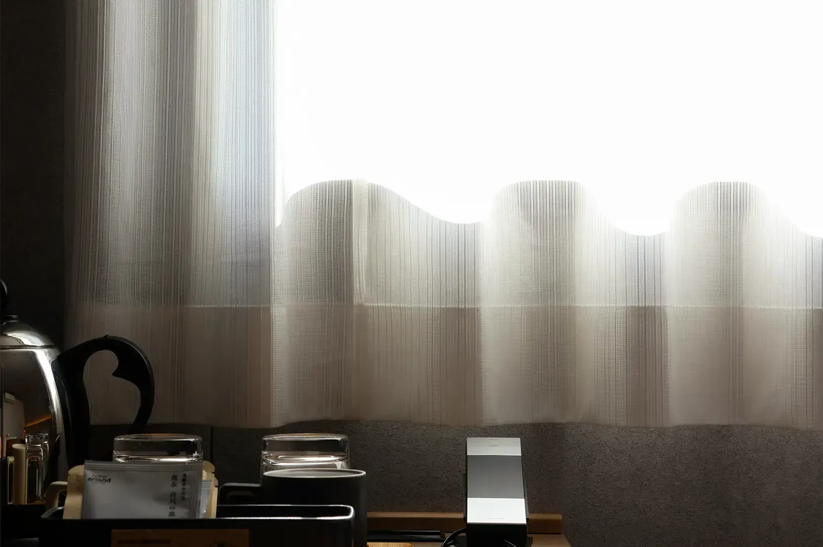Tea cups and kettle setup placed near a window with white curtains.