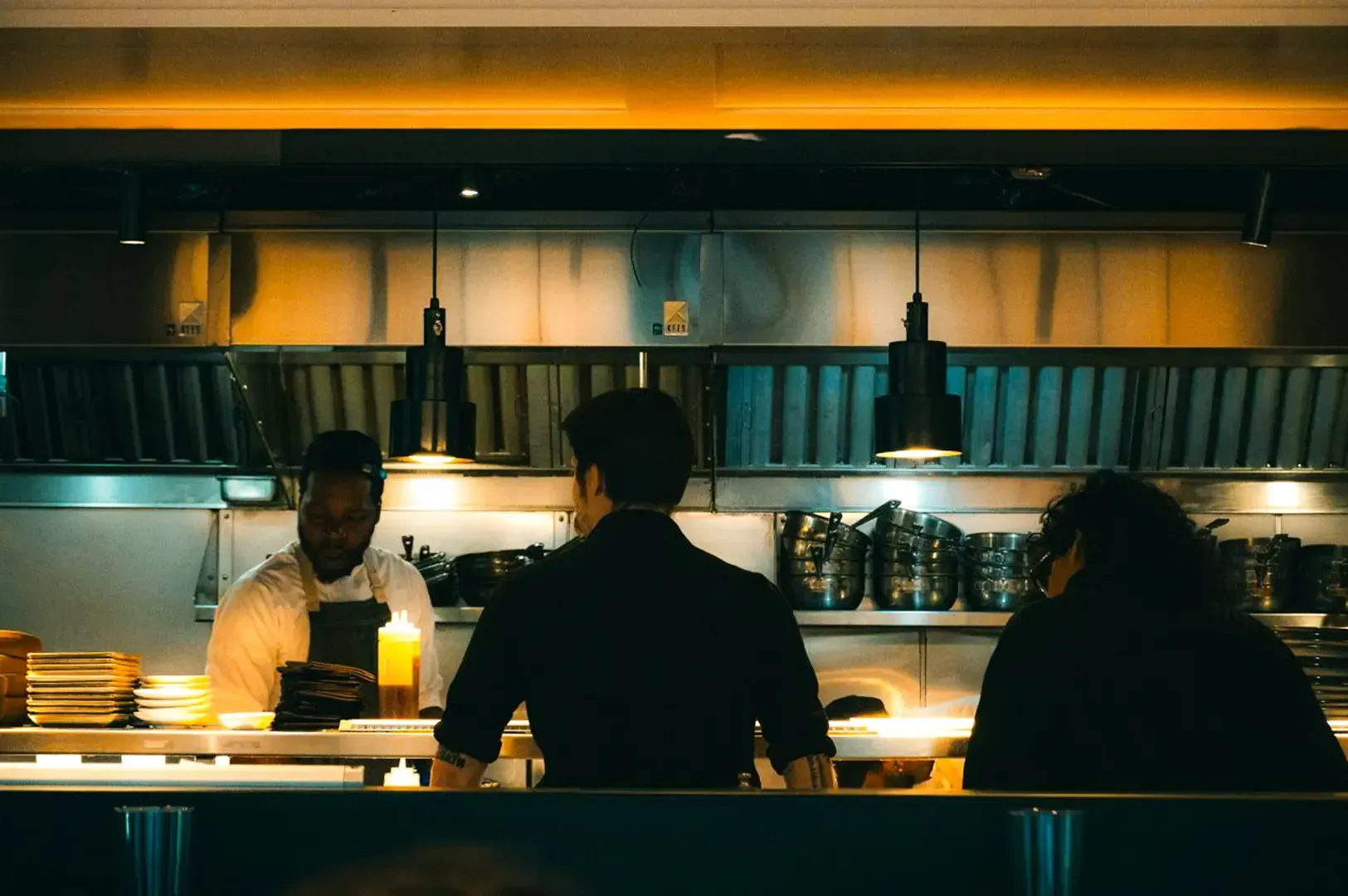 A dimly lit chef’s table with two customers waiting as the chef prepares a meal behind the counter.