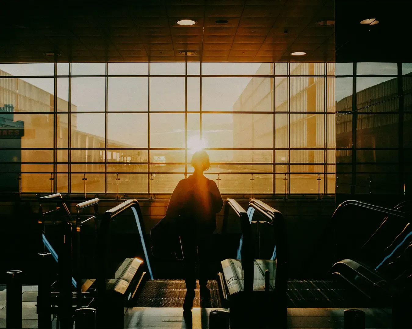 A woman slowly descending an escalator inside a modern airport terminal while natural light pours through large glass windows, creating a calm travel atmosphere.