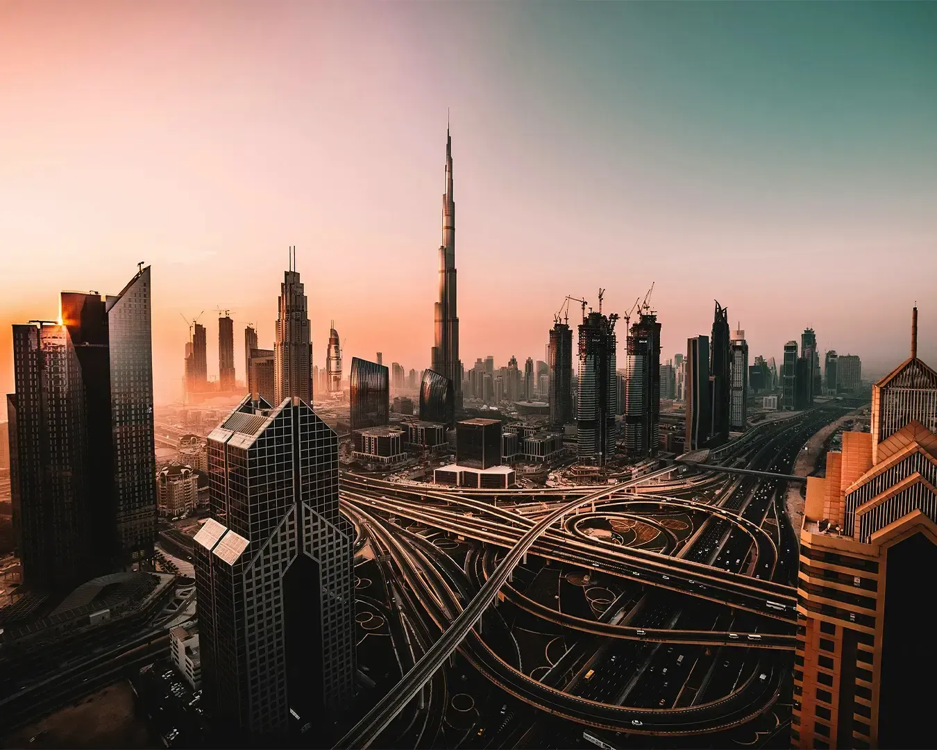 Aerial view of Dubai at sunset, showcasing the Burj Khalifa surrounded by modern skyscrapers and intricate highways, creating a dynamic urban skyline.