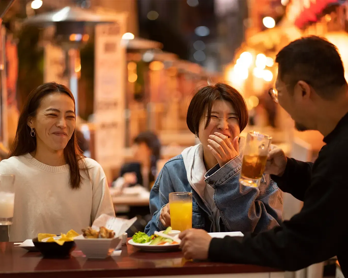 Three individuals seated around a table, smiling and engaged in conversation while sharing a meal, c