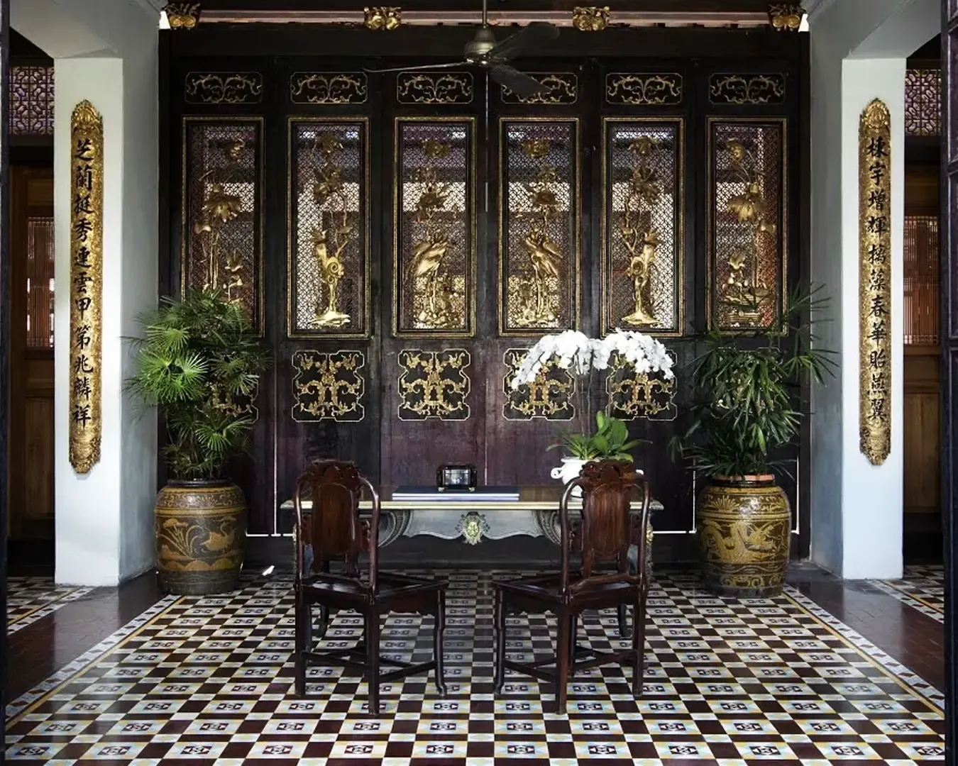 Elegant room with ornate wooden panels featuring gold accents and intricate carvings. Two wooden chairs face a table adorned with white orchids.