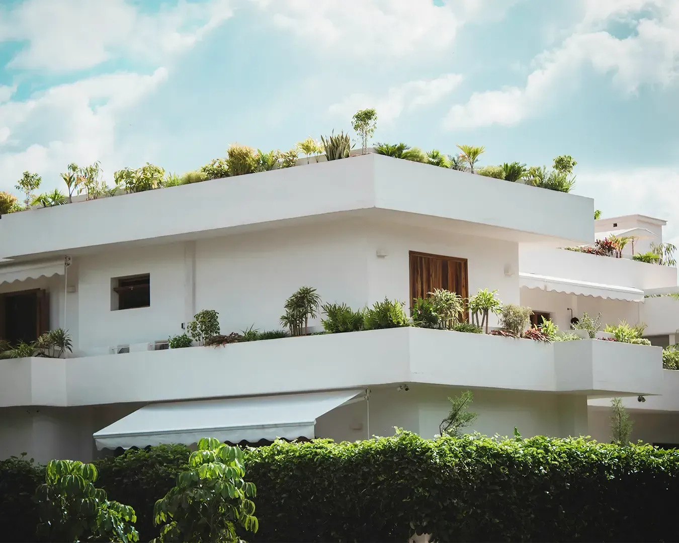 Modern white building with lush rooftop garden and balcony plants under a partly cloudy sky, conveying a serene and eco-friendly atmosphere.