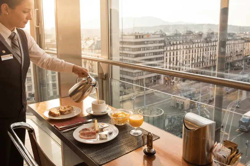 A server in a crisp uniform pours coffee into a white cup at a sunlit breakfast counter next to a la