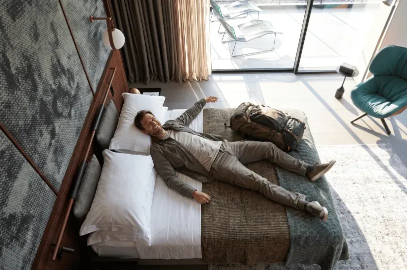 A high-angle view of a smiling man dressed in travel clothes collapse-lying starstruck across a hote