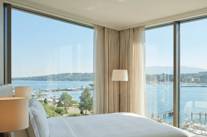 A pristine, neutral-toned hotel bedroom featuring a crisp white bed and expansive floor-to-ceiling w