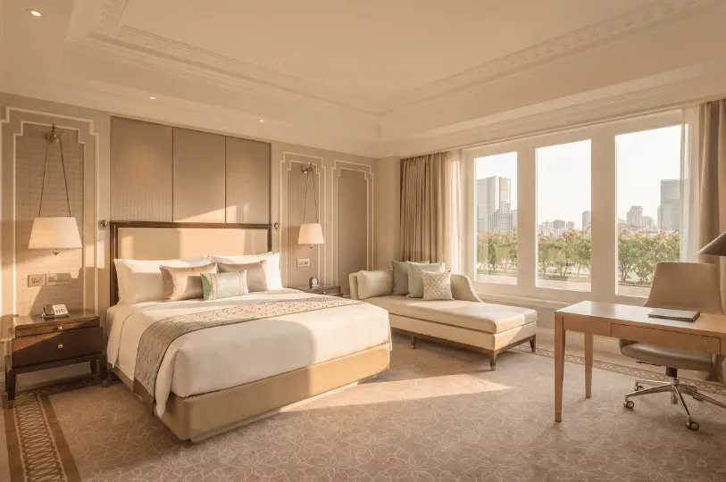 A sunlit hotel bedroom features a king bed on the left, a chaise lounge centered near large windows,
