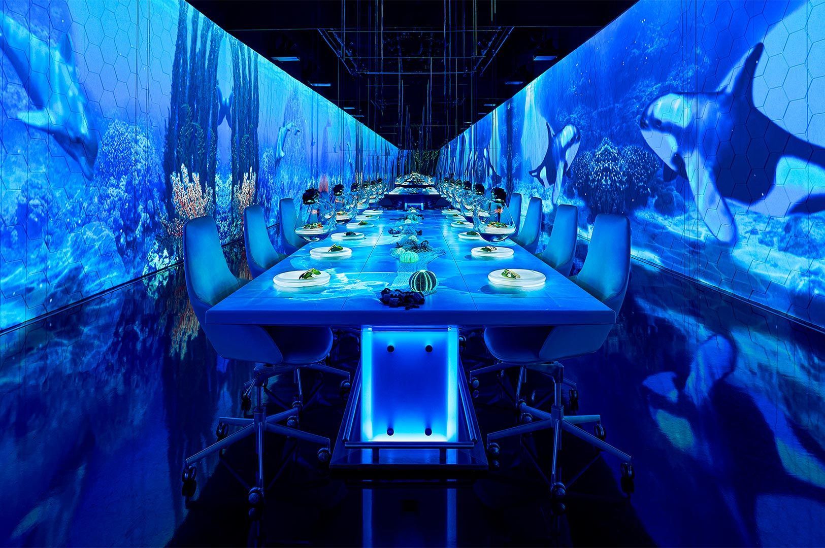 Long dining table in a high-tech restaurant with full-wall projection screens showing an immersive blue underwater scene with whales and coral.