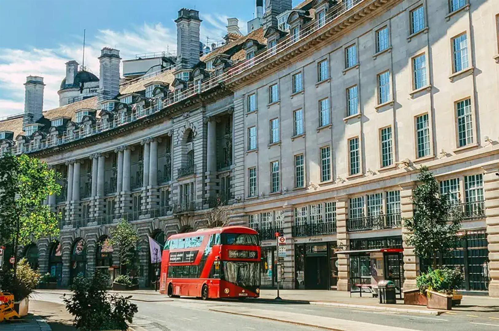 Red double-decker bus traveling along a quiet city street lined with historic stone buildings featur