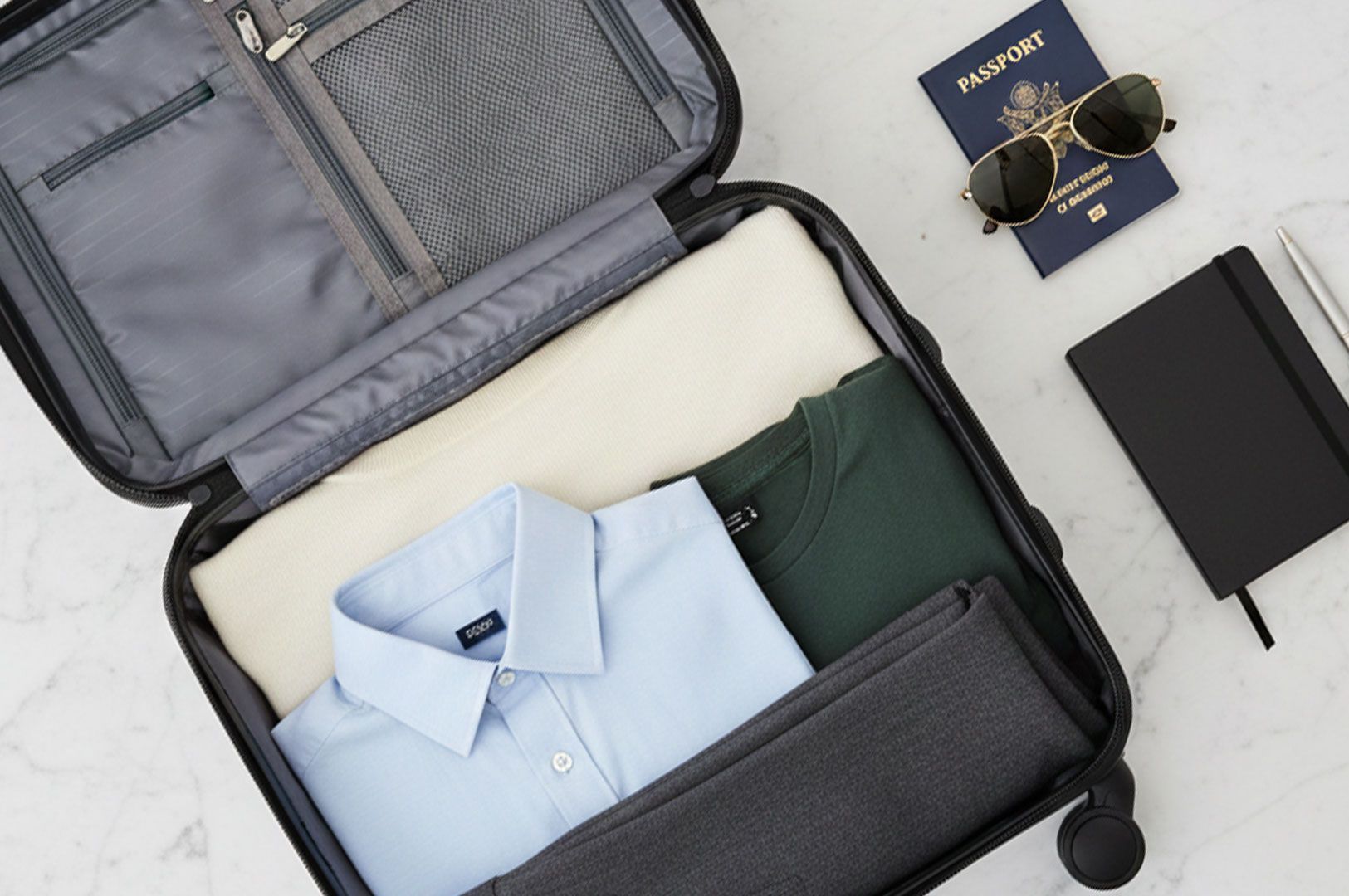 Top-down view of an open carry-on suitcase neatly packed with folded shirts and pants, placed next to a passport, aviator sunglasses, and a black notebook on a marble surface.