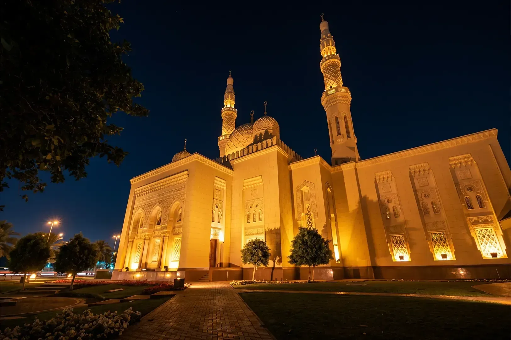 Famous religious spot Jumeriah Mosque in Dubai lit by its yellow warm lights and surrounded by greenery under a dawn sky.