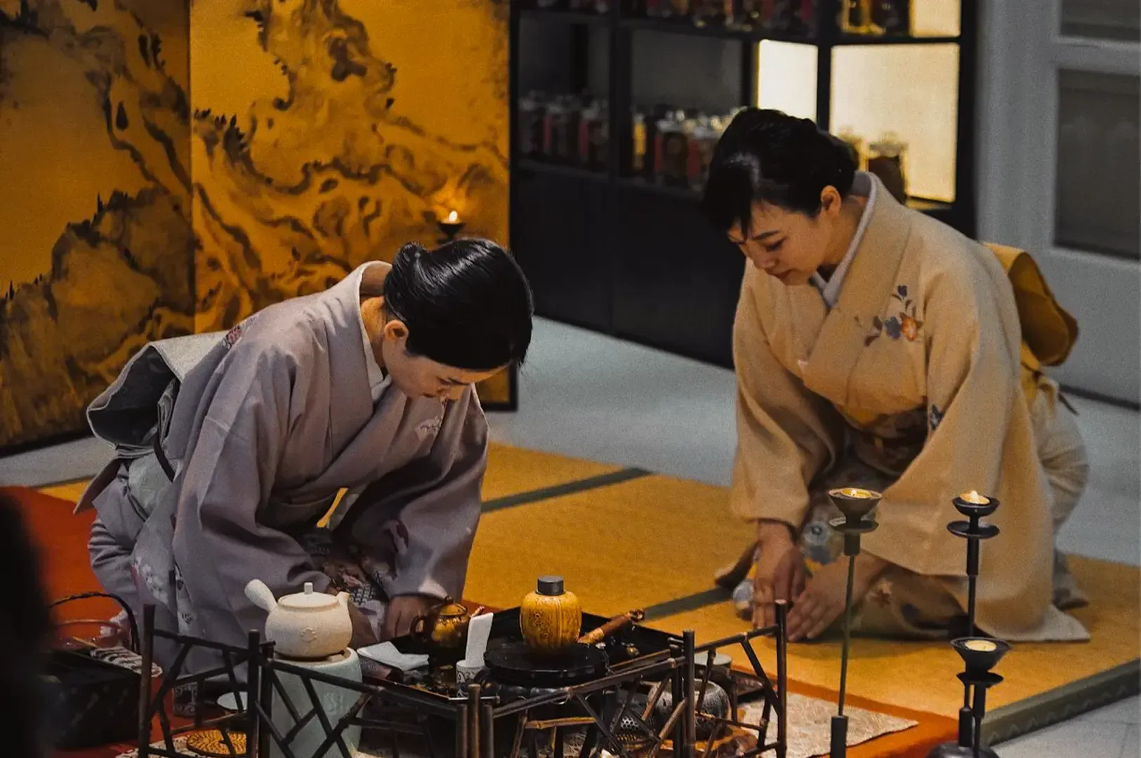 Two women in traditional Japanese clothing kneeling and bowing to each other to show respect with panels, trays, floor. and shelves in traditional Japanese style.